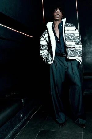 Man in patterned jacket posing in dark indoor setting, wearing button-down shirt and wide-leg trousers. Fashion editorial photography.