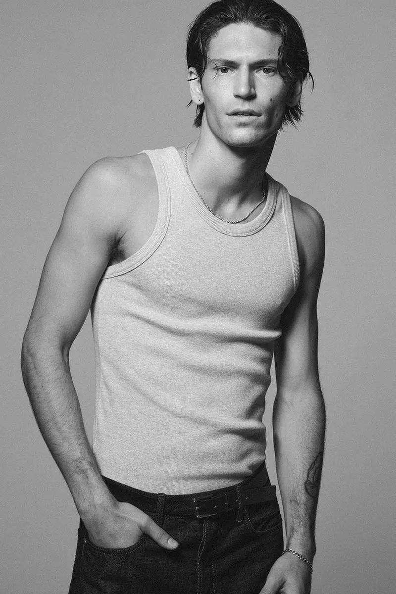 A young adult male model poses indoors wearing a tank top and jeans in a studio shot, showcasing fashion and lifestyle elements.