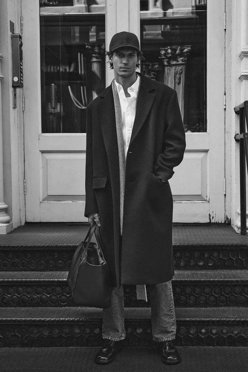 Monochrome portrait of a man in a coat, shirt, baseball cap, and jeans holding a bag, standing on steps against a building facade.
