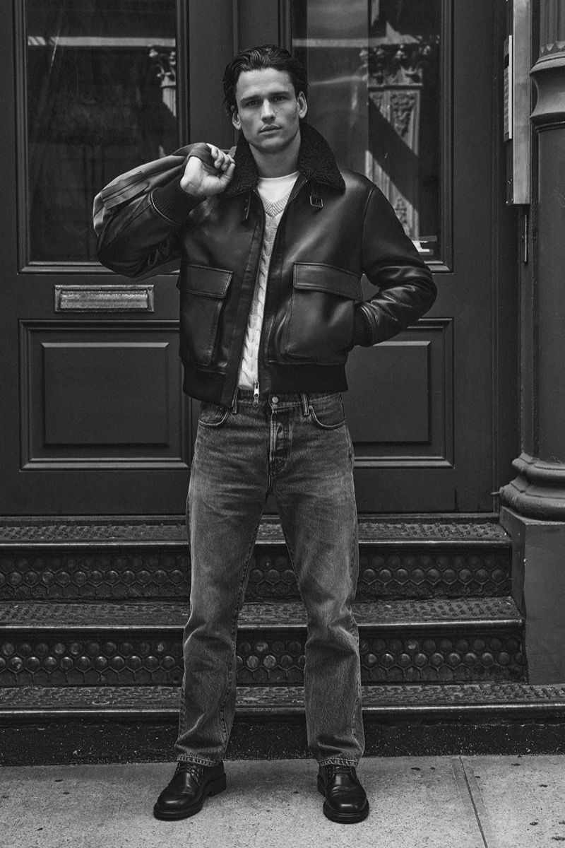 Monochrome portrait of a male model wearing a leather jacket standing on steps in front of a building exterior.