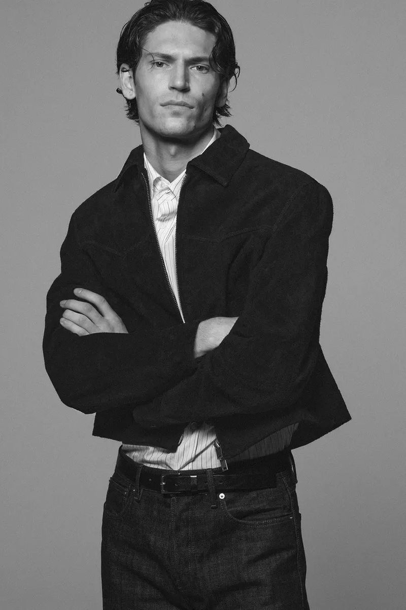Monochrome portrait of a male model wearing a dark jacket and collared shirt with arms crossed against a plain grey background