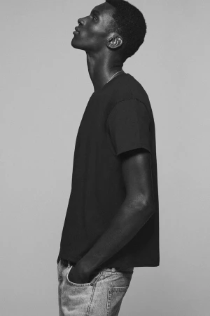 Black model wearing a dark t-shirt and jeans with hands in pockets in profile view against a grey background.
