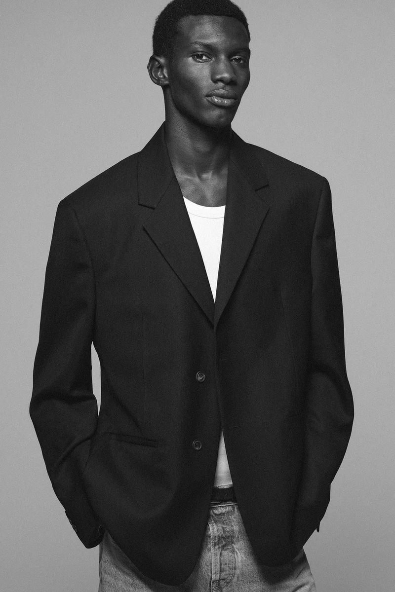 Black man wearing a dark oversized blazer over a white t-shirt and denim jeans against a solid grey background.