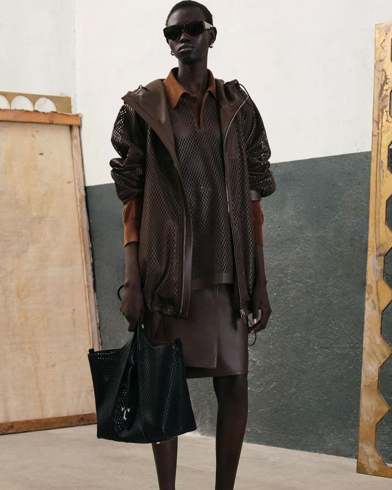 An adult female is posing indoors, modeling a dark brown leather skirt and a see-through mesh top and jacket, while carrying a black tote bag.
