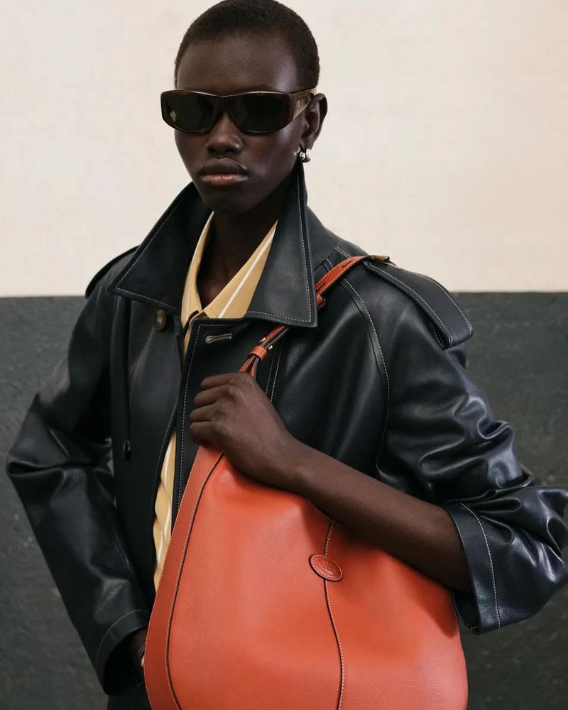 Dark-skinned woman models a black trench coat and orange tote bag in a fashion setting. Sunglasses and neutral background.