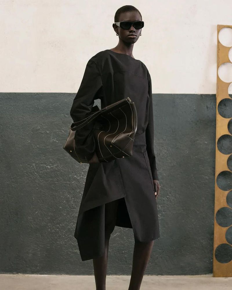 Fashion model presents black dress and tote bag in studio. Sunglasses, dark skin, modern design, and indoor lighting highlight the scene.