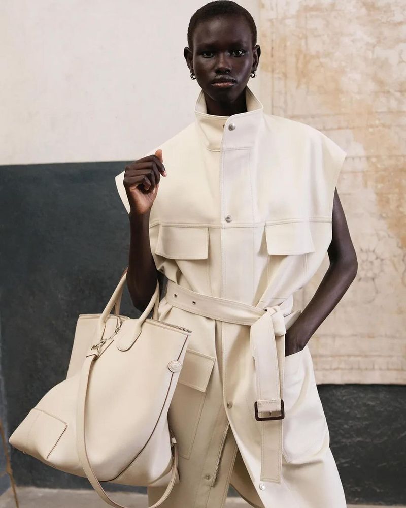 A dark-skinned model presents fashion, wearing a sleeveless cream-colored belted trench coat and carrying a matching tote bag against a neutral background.