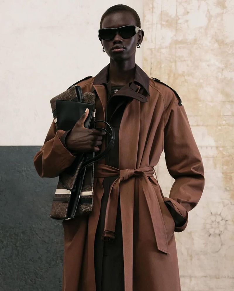 Model wearing a brown trench coat, black sunglasses, and carrying a bag, standing indoors against a textured wall.