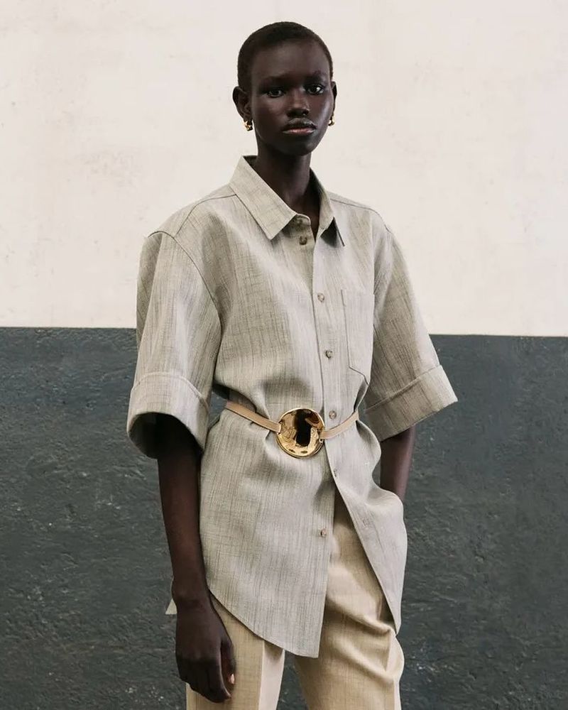 Fashion portrait of an African-descent woman wearing a beige button-down shirt, matching trousers, and gold belt, posing indoors.