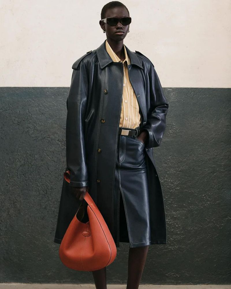 A woman poses in a blue leather trench coat and matching skirt, holding an orange leather hobo bag, showcasing a chic fashion ensemble.
