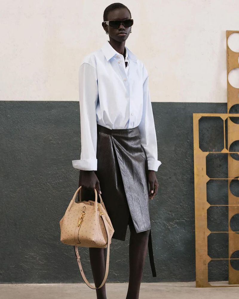 Dark-skinned woman with sunglasses wearing an unbuttoned white shirt and dark skirt, holding a beige handbag with a shoulder strap.