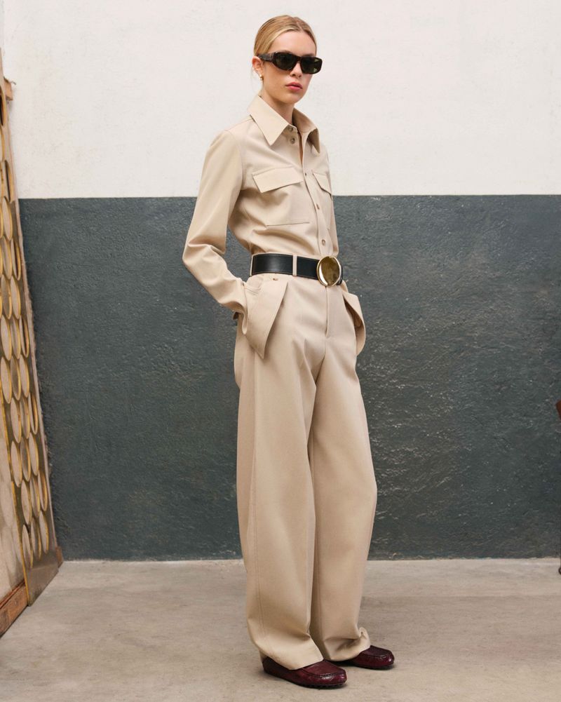 Woman posing in a beige shirt and wide-leg pants with a black belt, sunglasses, and maroon shoes in a studio with painted walls.