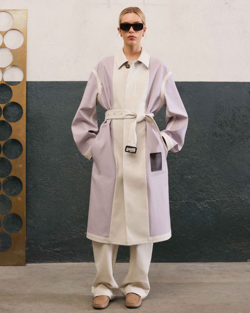 A fair-skinned adult fashion model in a color-blocked trench coat, loose pants, and sunglasses poses indoors in front of a two-tone wall.