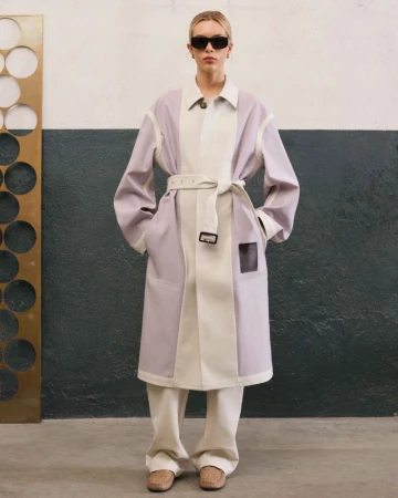 A fair-skinned adult fashion model in a color-blocked trench coat, loose pants, and sunglasses poses indoors in front of a two-tone wall.