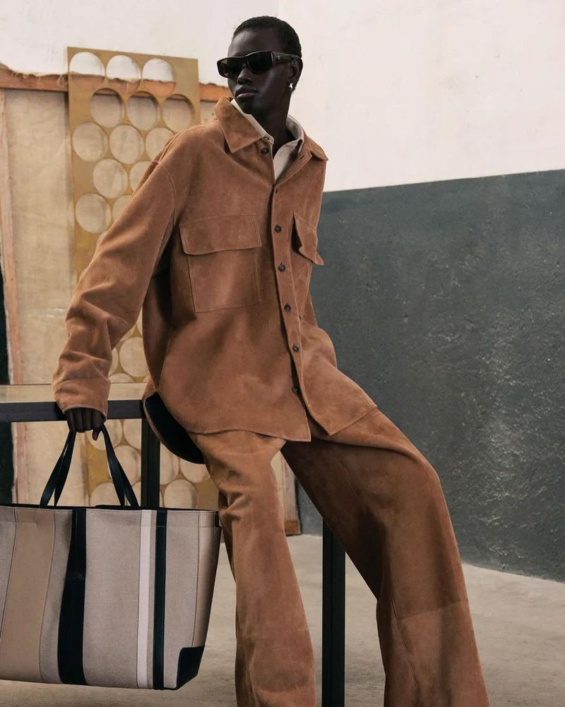 Man in tan suede suit and sunglasses poses with a tote bag against a neutral backdrop inside a studio, fashion editorial shot.