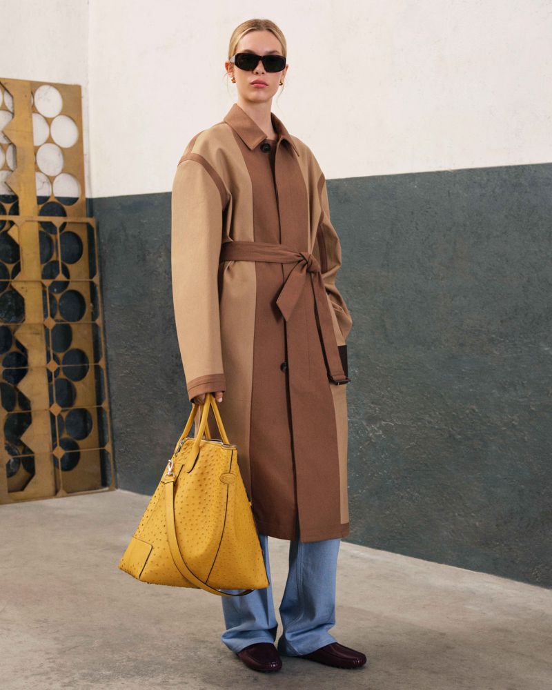 A fair-skinned woman in sunglasses, a two-tone trench coat, blue jeans, and flats, carrying a yellow leather handbag indoors.