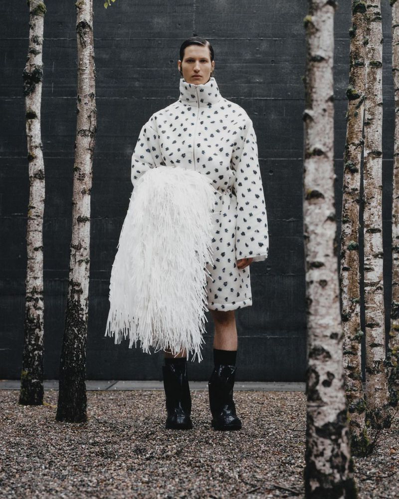A person in a spotted coat stands among birch trees, holding a fluffy accessory, wearing shiny black boots against a gray backdrop.