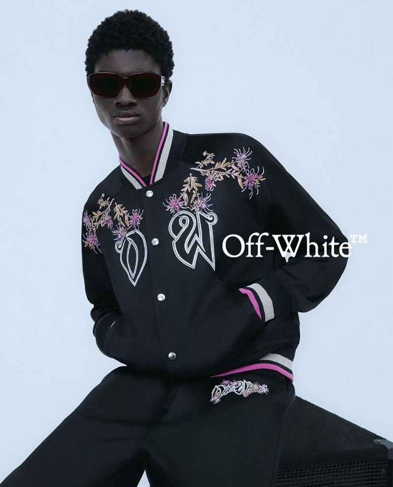 A model poses, wearing a black bomber jacket with floral embroidery and sunglasses, against a light blue backdrop and audio equipment.