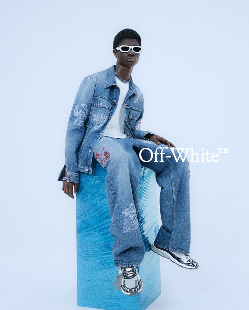 Man in denim jacket and jeans with white sunglasses sitting on a blue wrapped cube in a fashion shot against a light blue backdrop.