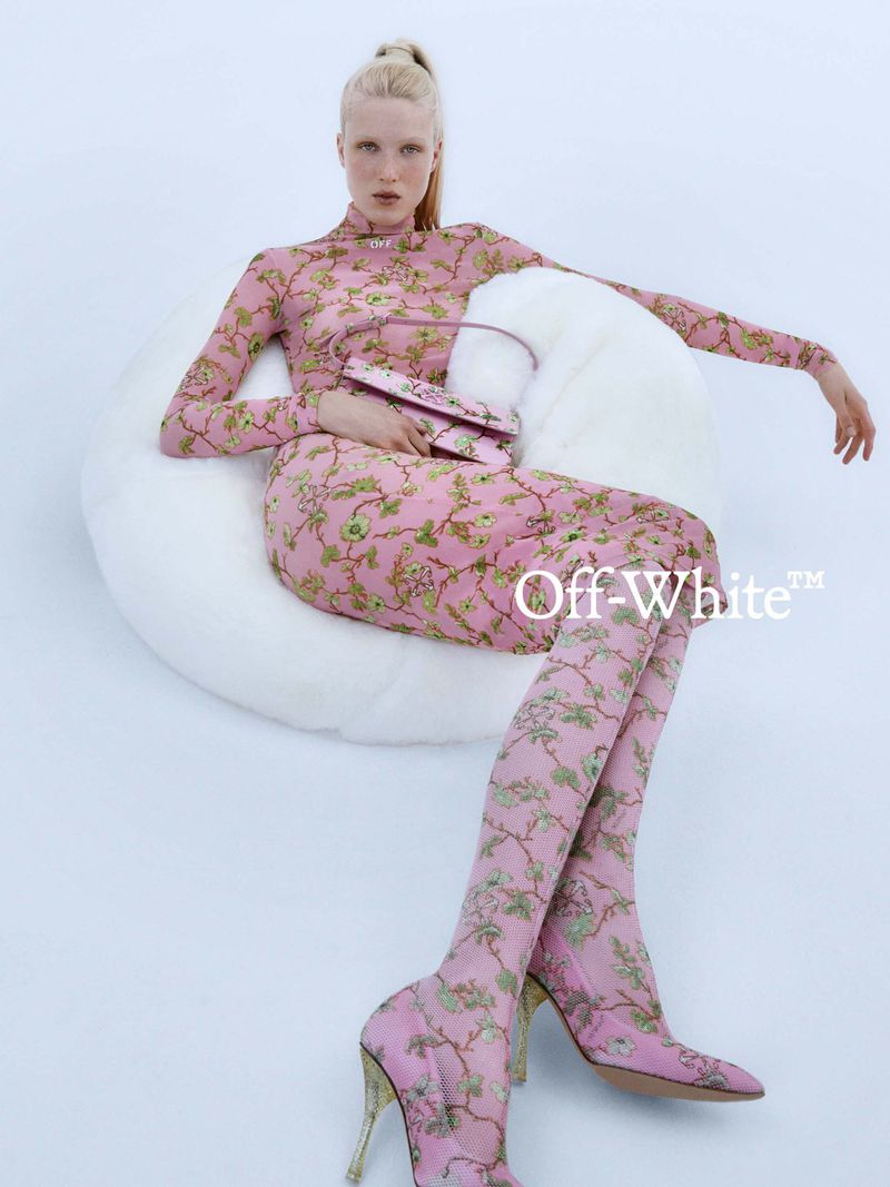 A woman in a pink floral body suit, skirt, and thigh-high boots poses on a white circular couch in a fashion studio shot.