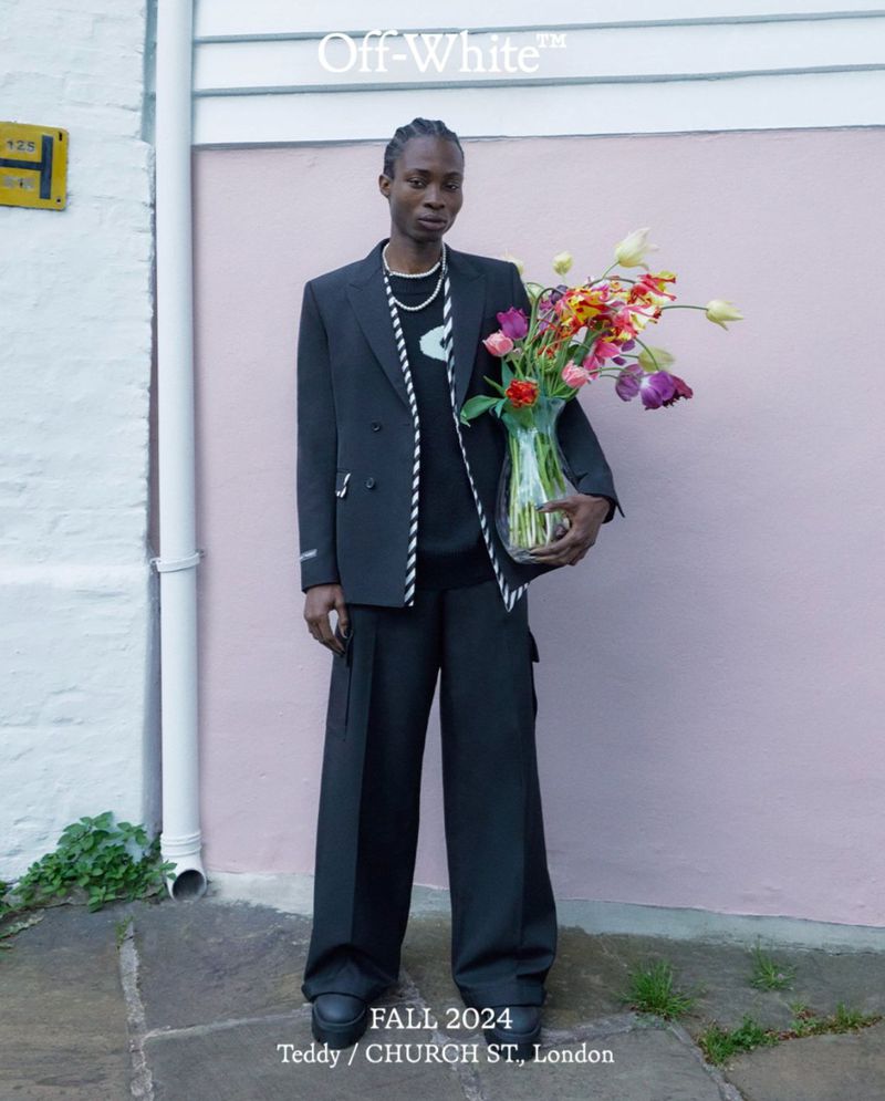 An adult male poses outdoors in a black suit with flowers, standing against a light pink wall with greenery and visible text overlay.