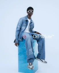 Model wearing a blue denim jacket and matching jeans seated on a bright blue wrapped block against a pale studio background.