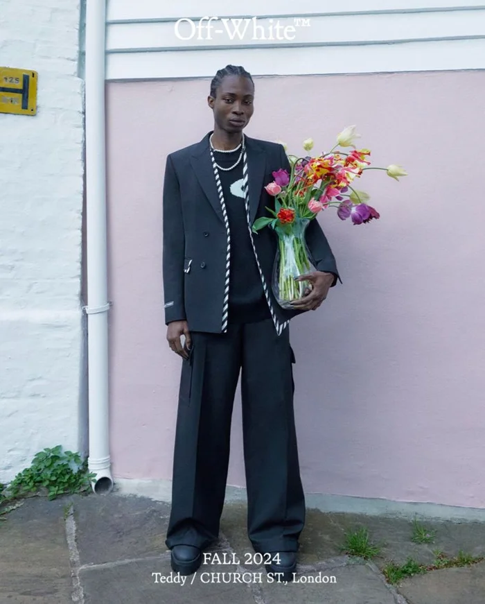 Model wearing a black blazer and wide trousers holding a glass vase of colourful flowers against a pink wall