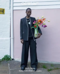 Model wearing a black blazer and wide trousers holding a glass vase of colourful flowers against a pink wall