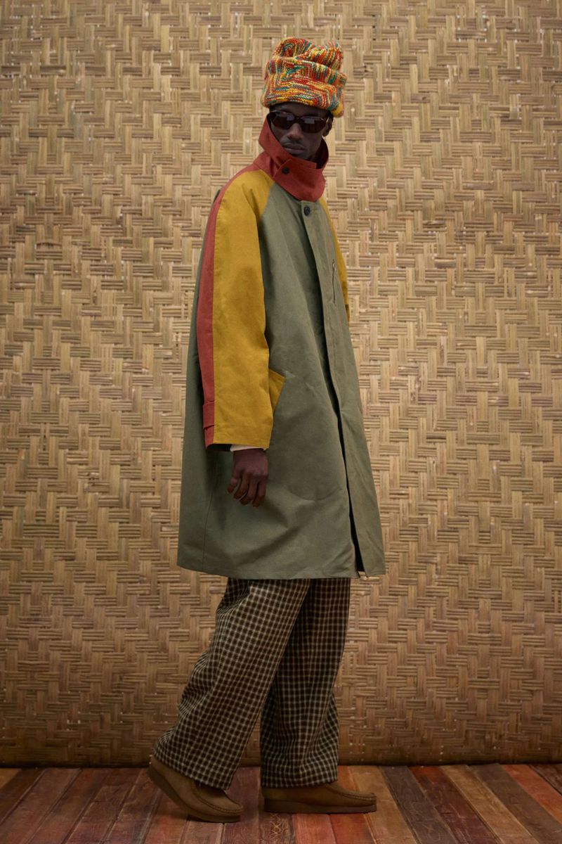 An adult Black male fashion model in a hat, coat, and patterned pants poses in a studio with a woven background.