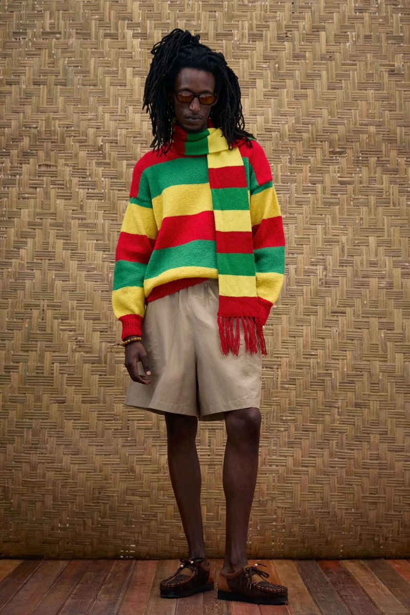 Man in Rasta-colored knit sweater and shorts posing indoors against woven fiber wall with a serious facial expression and stylish glasses.