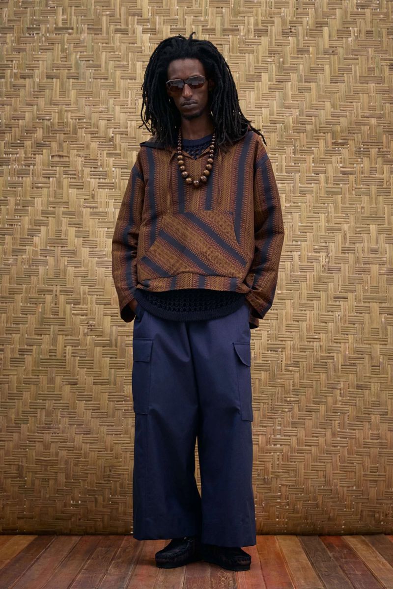 Full-length shot of African-American male model wearing a brown striped hooded shirt and navy wide-leg pants, standing indoors.