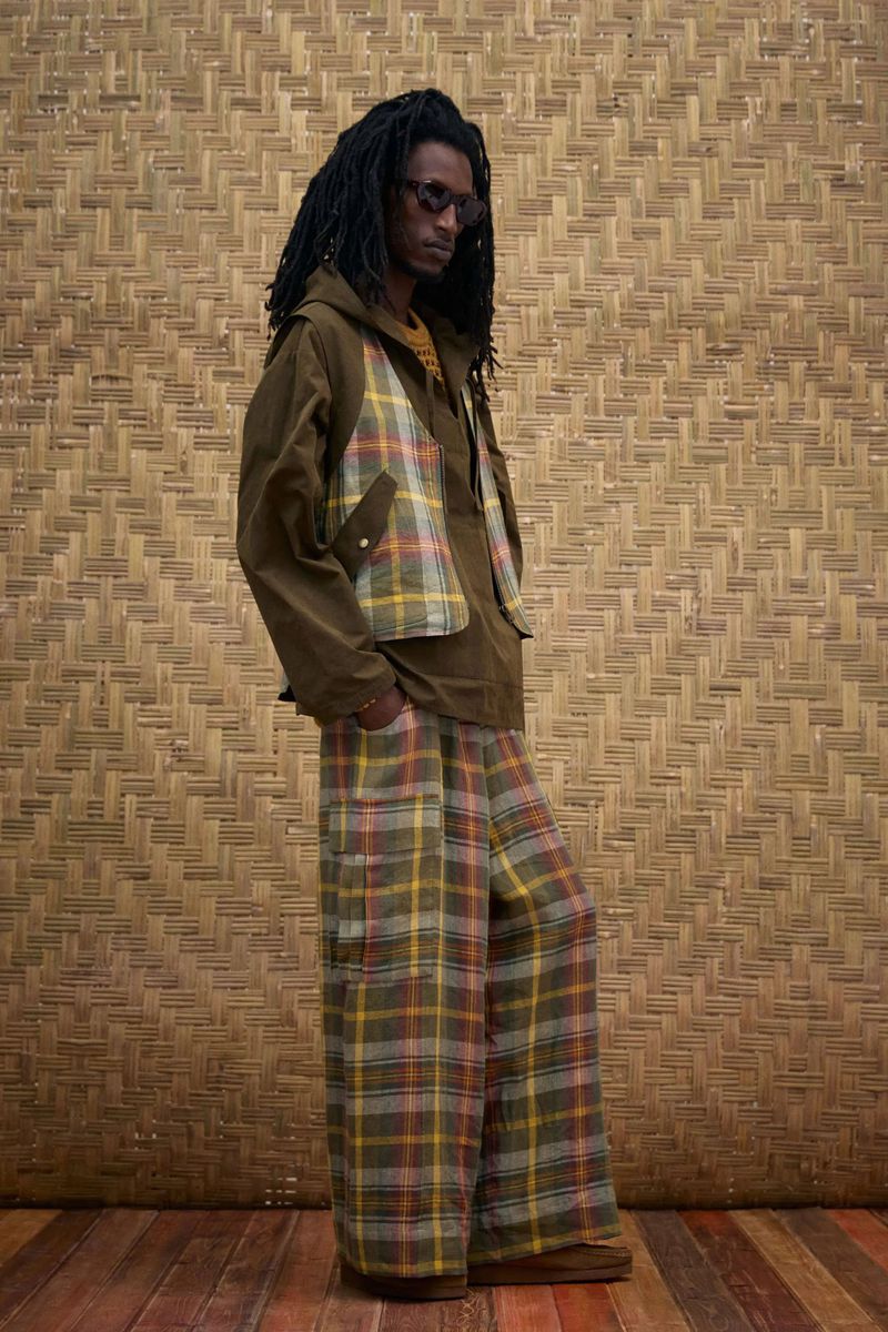 A man in sunglasses poses in an olive jacket with a plaid vest and matching plaid pants against a woven bamboo background.
