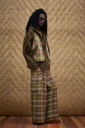 Model with long dark hair wearing a patterned plaid vest and trousers against a woven wall