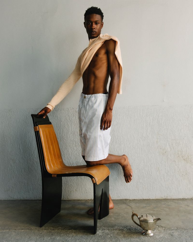 A slender, athletic Black man poses on a chair in a studio, wearing shorts and a sweater draped over his shoulder. Silver teapot visible.