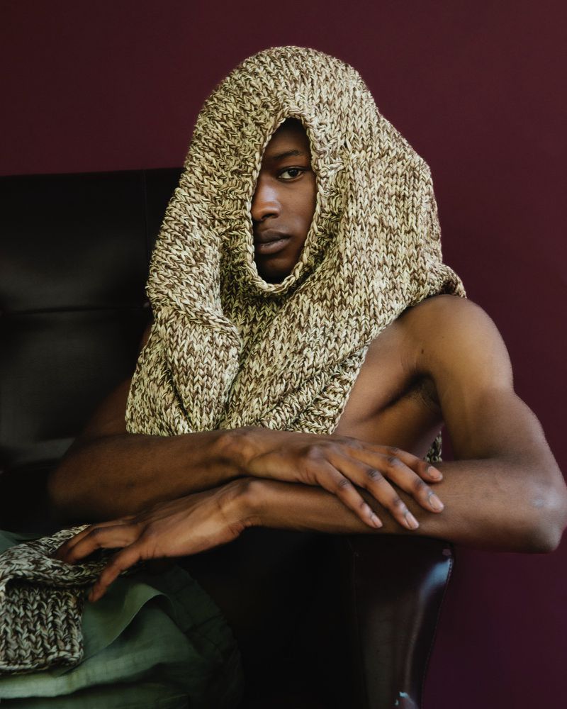 Portrait of an African American adult male with knit hood. He sits in a dark chair with his arms folded in an indoor studio.