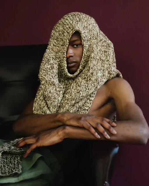 Portrait of an African American adult male with knit hood. He sits in a dark chair with his arms folded in an indoor studio.
