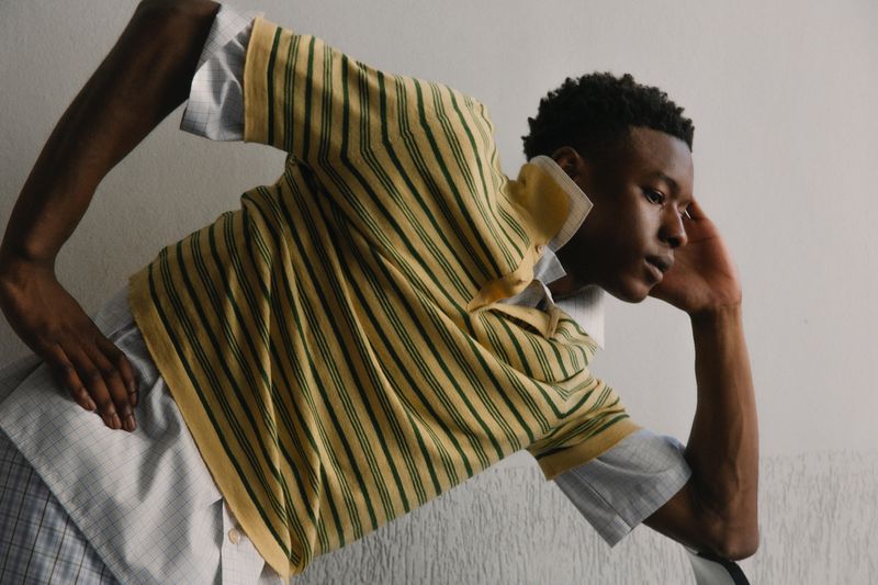 Young adult with dark hair poses indoors wearing a striped yellow polo shirt with a grey patterned shirt underneath in a fashion shot.
