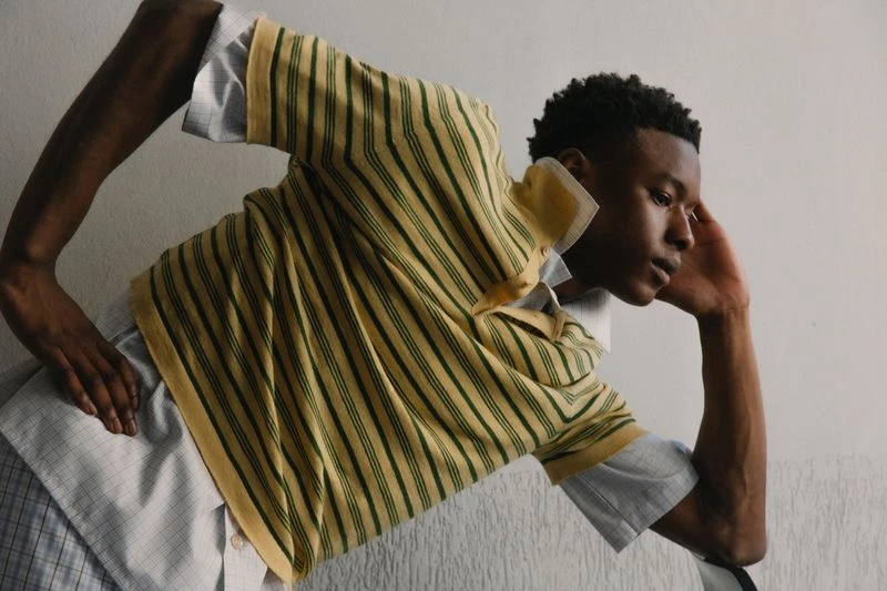 Young adult with dark hair poses indoors wearing a striped yellow polo shirt with a grey patterned shirt underneath in a fashion shot.