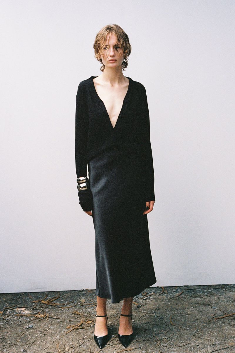 A slender adult woman poses in a black sweater dress and black high heels against a plain, light-colored wall indoors.