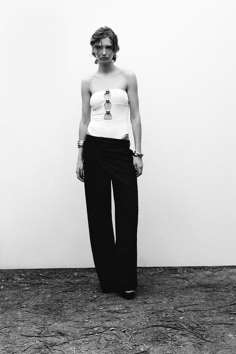 A woman in a strapless top and wide-leg pants stands against a stark white wall, creating a minimalist fashion portrait in monochrome.