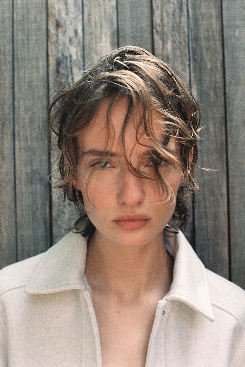 Close-up portrait of a fair-skinned adult woman with shoulder-length brown hair and a light-colored jacket against a wooden wall.