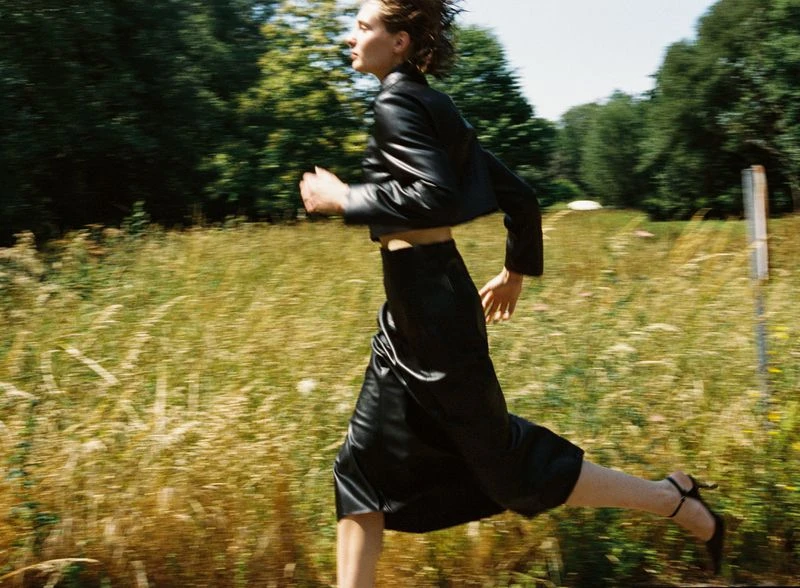 Woman in black leather skirt and jacket is running across a grassy field with trees in the background, Fashion photography.