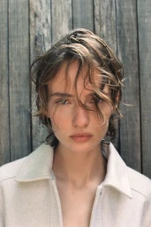 Portrait of a model with tousled wet hair wearing a light-coloured collared jacket against a vertical wooden slat wall