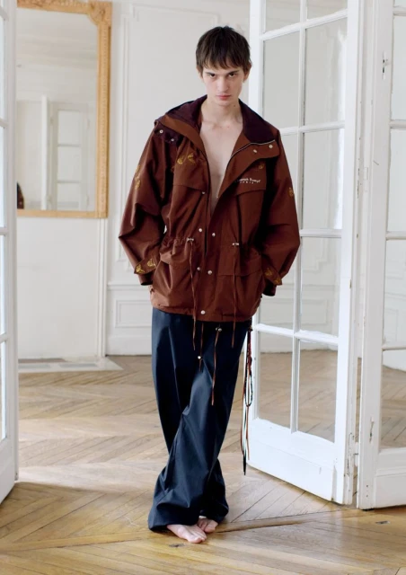 A young man with shaggy brown hair poses barefoot in an open brown hooded jacket and dark pants, standing in front of white doors.