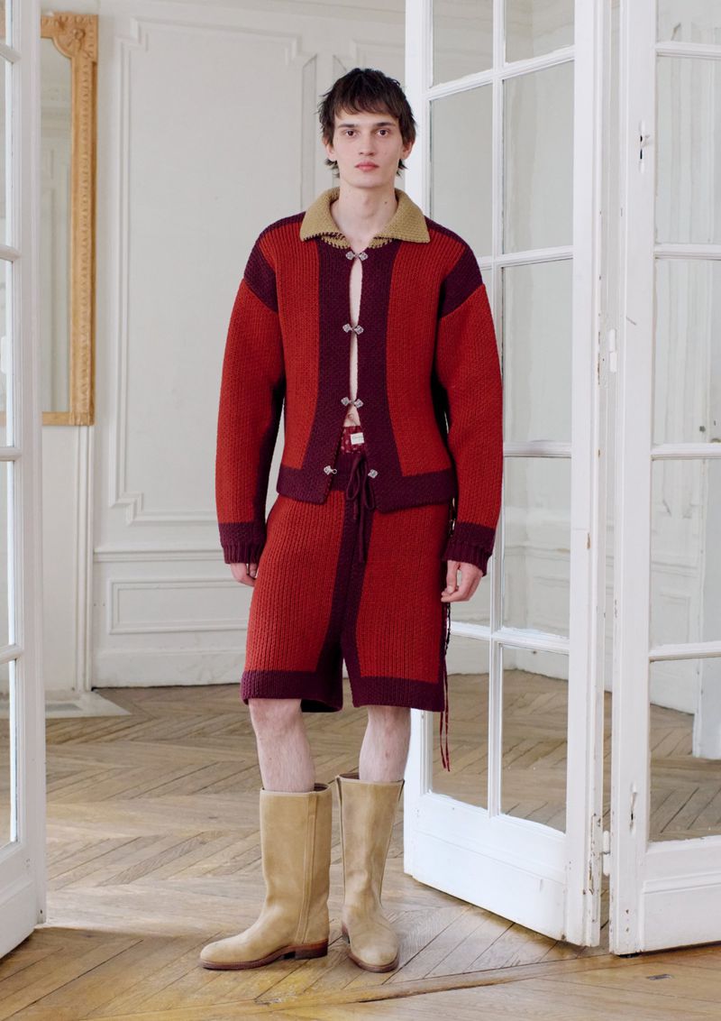 Male fashion model wears a knit cardigan and matching shorts with high boots, posing indoors near a white paneled door and decorative mirror.