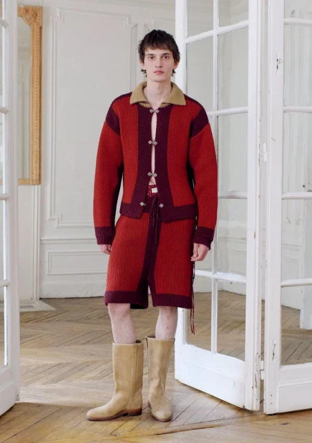 Male fashion model wears a knit cardigan and matching shorts with high boots, posing indoors near a white paneled door and decorative mirror.