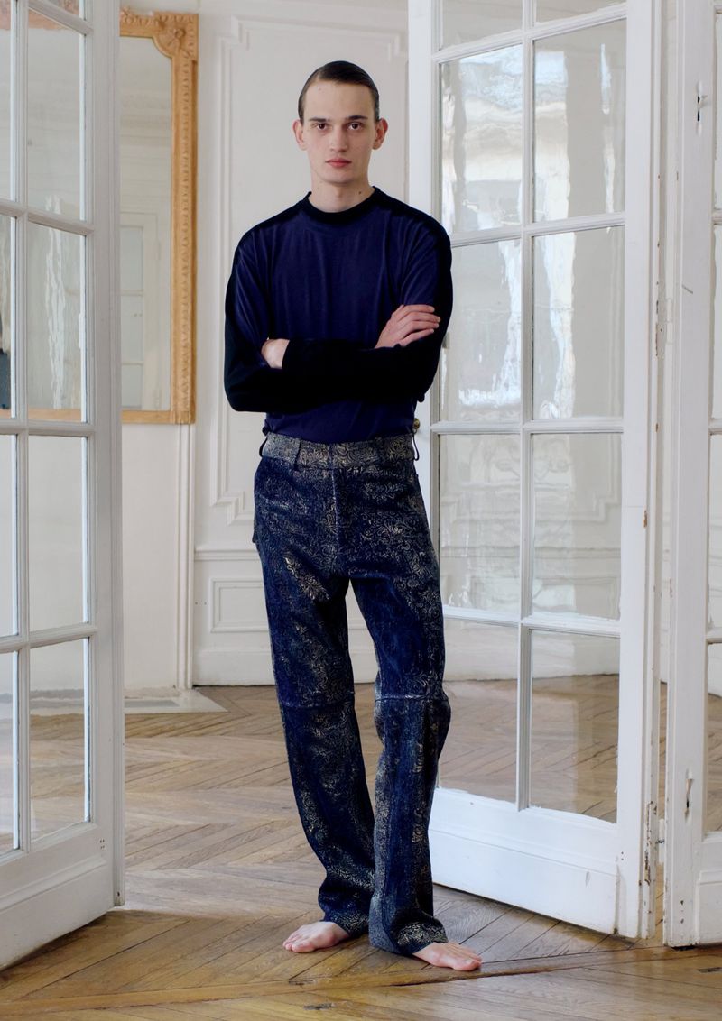 A slender, fair-skinned man with a slight frown stands barefoot in a doorway, wearing a long-sleeved blue top and mottled blue-gray pants.