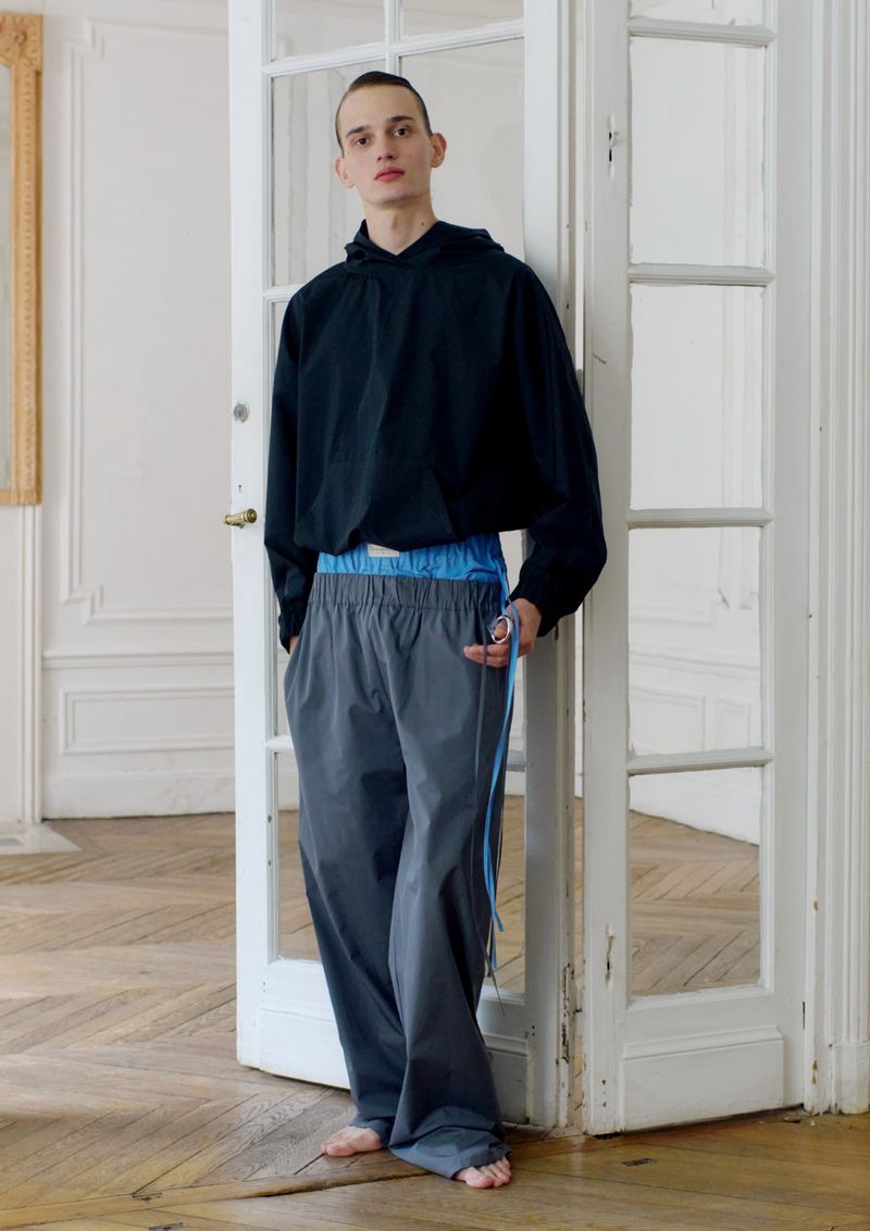 Male fashion model poses indoors wearing a black hoodie and grey sweatpants with a blue waistband and side stripe, leaning in doorway.