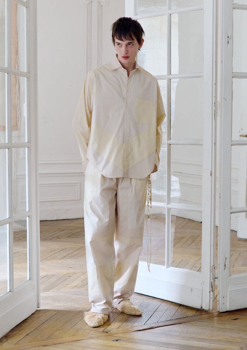 Person in relaxed pose wearing light-colored, loose-fitting shirt and pants, standing in a room with white-frame double doors.