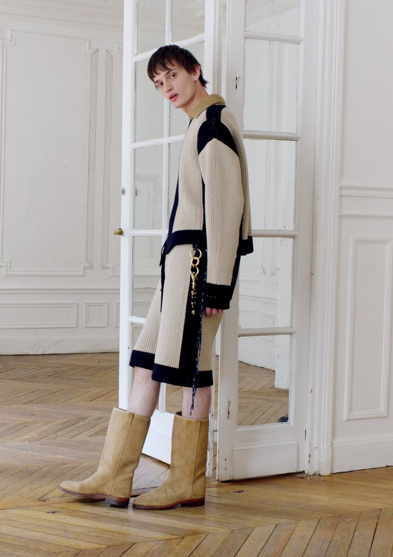 Male model wears an ivory and navy jacket with matching shorts, posing in a minimalist interior near a white door frame.